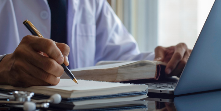 Closeup male doctor or physician reading text book and writing information,work on laptop computer with medical stethoscope on t