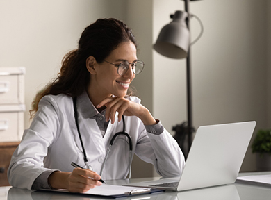 Smiling professional female doctor taking notes, looking at laptop